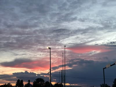 The image captures a dramatic sunset over Laagri alevik, Estonia, with vibrant pink and orange hues illuminating the clouds. Several streetlights are visible, some with their lamps illuminated, casting a soft glow. A security camera is attached to one of the lampposts. In the distance, trees line the horizon, and power lines crisscross the sky. A traffic sign is also present, indicating directions. The overall scene is one of a serene evening, with the natural beauty of the sunset contrasting with the man-made elements of the urban landscape.