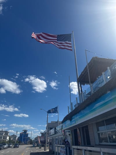 The image captures a sunny day in Hampton, United States of America, likely along the coast. A large American flag, proudly displayed on a flagpole, billows in the breeze against a vibrant blue sky dotted with fluffy white clouds. To the right, a building with a thatched roof and a balcony overlooking the street is visible. The building appears to be a commercial establishment, possibly a restaurant or shop, with signage that includes the numbers "1923" and "1925". Further down the street, other buildings and a blue, multi-story structure under construction are visible. A second flag, featuring a blue field with a seal, flies from a lamppost. A road with cars driving on it runs alongside the buildings. People are visible on the sidewalk and on the balcony of the building with the thatched roof. The overall atmosphere suggests a lively, seaside town setting.