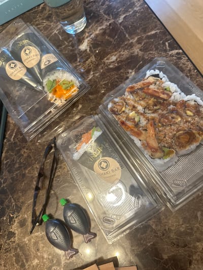 This image captures an indoor scene on a dark brown, polished marble-patterned surface, located in Vandœuvres, Switzerland. The primary subjects are several clear plastic takeout containers holding sushi.
Specifically, two rectangular, clear plastic containers are stacked in the upper left, each containing a single temaki (hand-rolled sushi cone). Both display circular labels reading "KINUM TEMAKI BAR BY KAKINUMA," with one additionally marked "100% RECYCLABLE." The visible temaki features white rice, orange roe, and green ingredients. To the right, a larger, clear plastic container holds multiple sushi rolls (maki) topped with a dark sauce, pieces of cooked shrimp or tempura, and avocado. Another empty or partially visible temaki container, identical to the stacked ones, is positioned below the larger maki container.
Adjacent to the containers, a pair of dark-framed eyeglasses lies on the surface. To their right are two black, fish-shaped plastic containers with bright green caps, commonly used for soy sauce. A clear glass filled with water is visible in the upper center of the frame. In the upper left background, partial light blue or white text, "LUCITE," is visible on a lighter surface. No individuals are present.