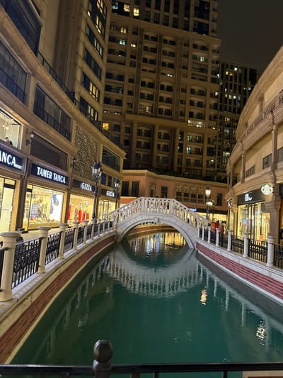 An outdoor, nighttime scene depicts a Venetian-themed commercial and residential complex located in Esenler, Turkey. A central, artificial canal with dark green water flows from the foreground towards the background, spanned by a white arched pedestrian bridge with decorative balustrades. The water surface reflects the surrounding artificial lights and architectural elements.

Two multi-story buildings flank the canal. The building on the left features ground-level storefronts including "TAMER TANCA" and "GIZIA," exhibiting merchandise through large glass windows. Above these shops, higher floors display illuminated windows, and a discernible individual with a dark face covering is visible inside an upper-level space. The building on the right also hosts ground-level businesses, including a visible Starbucks logo, and shows a small cluster of indistinct individuals in front of its brightly lit storefronts. Both buildings exhibit classical architectural elements such as arches, columns, and patterned facades, with higher floors displaying multiple illuminated windows against the dark night sky.

Decorative black metal railings with balusters line both banks of the canal, featuring integrated ornate black streetlights with glowing globes. Red brickwork forms the base of the canal walls. In the background, taller, modern high-rise buildings with numerous illuminated windows rise against the dark sky, suggesting a dense urban environment. No specific actions or interactions beyond general pedestrian presence are clearly discernible.