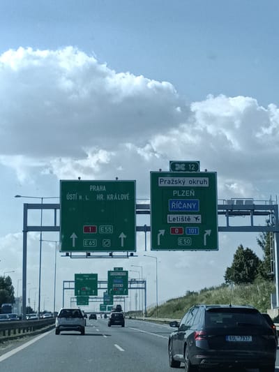 This media file depicts a view from a car driving on a highway in the Czech Republic, likely near Říčany. The image focuses on overhead highway signs and the road ahead.
The main subjects are the green highway signs, which are clear and legible. The left sign indicates "PRAHA" (Prague), "ÚSTÍ N. L." (Ústí nad Labem), and "HR. KRÁLOVÉ" (Hradec Králové), with route numbers 1, E65, and E55. The right sign displays "Pražský okruh" (Prague Ring Road), "PLZEŇ" (Plzeň), "ŘÍČANY" (Říčany), and "Letiště" (Airport), with route numbers 12, 0, 101, and E50.
The scene is a multi-lane highway with other vehicles visible, including cars and a truck. The weather appears to be clear and sunny, with bright blue sky and fluffy white clouds. The time of day seems to be daytime, based on the lighting. The setting is a highway with grassy embankments and trees visible on the right side of the road.
Notable details include the license plate of the black car in the foreground, which reads "6SL 7937". The signs are typical for European highways, using green backgrounds and white lettering for destinations and route numbers. The presence of "Pražský okruh" and "ŘÍČANY" confirms the likely location. No specific emotions are conveyed, as the image is a still shot of a highway scene.