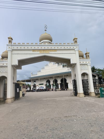 The image captures the grand entrance of the Mesjid Al-Ihsan Kemukiman Meuneubuk Grong-Grong in Grong-Grong, Indonesia. A prominent, ornate cream-colored archway forms the main gateway, featuring decorative pillars, intricate carvings, and small golden-domed minaret-like structures. A central dome on the archway is topped with a crescent moon and star symbol. A golden sign across the arch clearly displays the mosque's name: "MESJID AL-IHSAN KEMUKIMAN MEUNEUBUK GRONG-GRONG".

Beyond the open black metal gates of the archway, the main mosque building is visible, constructed in a similar light-colored style with multiple arched windows and a green trim along its roofline. Several motorbikes are parked in the paved area in front of the mosque, indicating visitors or congregants. The foreground shows a gravel pathway leading up to the entrance.

The scene is set outdoors during the daytime, under an overcast sky. Power lines are visible stretching across the top of the frame. Palm trees and other greenery can be seen in the background behind the mosque complex. No individuals are distinctly visible, but the parked motorbikes suggest a regular day of activity at the mosque.