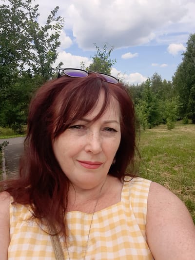 A woman with auburn hair and freckles, wearing a yellow and white checkered dress and sunglasses on her head, takes a selfie in a park. The background consists of trees, a paved path, and a cloudy sky. The image appears to be taken outdoors in a natural setting.