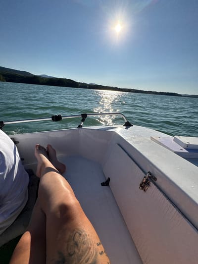 A first-person perspective shot shows a person's legs and feet propped up on the edge of a white boat sailing on a lake. The sun is bright and high in the sky, casting a strong reflection on the water. The water is a vibrant blue-green color, and gentle waves are visible. In the background, a tree-lined hill slopes down to the water, with a clear blue sky above. The boat appears to be on the lake in the vicinity of Golyama Zhelyazna, Bulgaria, judging by the surrounding landscape.