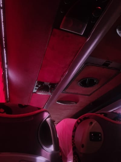 The image displays the interior of a passenger transport vehicle, likely a bus or coach, illuminated by a dominant red/magenta ambient light source, possibly LED strips visible on the left ceiling edge. The perspective is from a low angle, looking forward and slightly upwards. The ceiling features dark red paneled sections, interspersed with overhead storage compartments, circular air vents, and rectangular light fixtures. In the foreground, the upper portions of two dark-colored passenger seats are visible, framing the lower half of the scene. Between these seats, a single individual is seated further forward, with only the back of their head and shoulders discernible. This individual is wearing a bright pink head covering. The exact location within Al-Kiswa, Syria, cannot be determined from the visual evidence presented in the image.