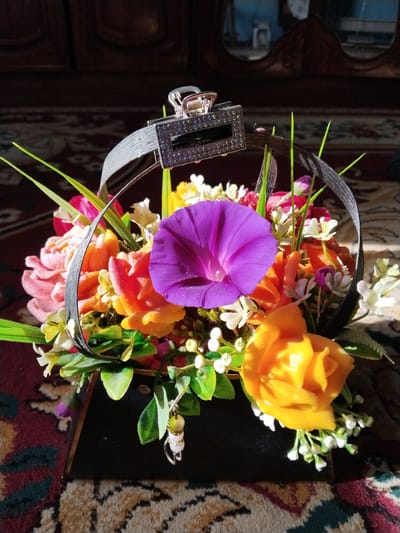 The image displays a decorative arrangement of artificial flowers in a dark, rectangular container, possibly a gift box or basket. The flowers are a mix of vibrant colors, including pink, orange, yellow, and purple, with some smaller white blossoms and green foliage. A black ribbon forms an arch over the arrangement, secured at the top by a silver hair clip embellished with small, dark stones. A small, dangling charm with beads is attached to the side of the container. The arrangement is placed on a patterned carpet with shades of red, green, and beige. The background consists of dark wooden furniture and a reflective surface, possibly a mirror or glass, showing a blurred interior scene. The lighting suggests an indoor environment with sunlight streaming in, creating shadows on the carpet. There are no people or animals visible in the image. The scene appears to be a still life, likely in a domestic setting. The location is Tinchlik, Uzbekistan.