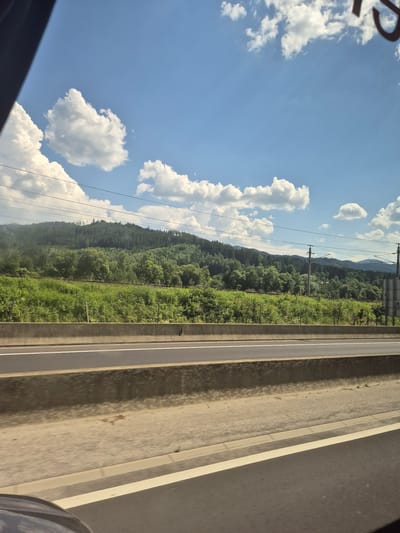 This image captures a scenic view from a vehicle traveling on a highway in Kindberg, Austria. The foreground is dominated by the highway, with multiple lanes visible and a concrete barrier separating them. Beyond the highway, a vibrant green landscape unfolds, characterized by lush fields and densely wooded hillsides. The hills rise towards the horizon, covered in a carpet of trees. The sky above is a brilliant blue, dotted with fluffy white cumulus clouds. Electrical power lines and poles are visible against the sky, indicating the presence of infrastructure near the road. The overall impression is one of a sunny day in a rural, mountainous region.