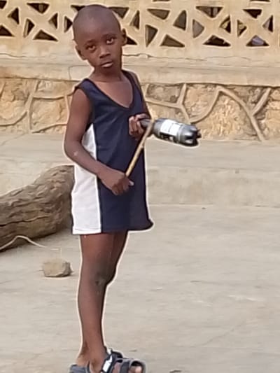 A young, slender boy stands outdoors, holding a stick attached to a plastic bottle. He wears a dark blue and white sleeveless dress with sandals on his feet. His gaze is directed forward, conveying a somewhat serious expression. A wall with a unique patterned design sits behind him. A log rests to his left, with a small rock nearby. The ground is a concrete surface.