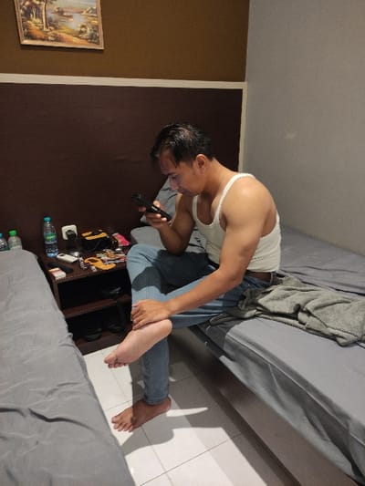 A young man wearing a white tank top and blue jeans is sitting on a bed. He is holding a black phone in his right hand and looking at it. His left hand is resting on his right thigh. His right foot is bare and on the floor. The bed is covered in a grey blanket. There is a wooden bedside table with a bottle of water and other items. The floor is tiled. The room is plain with brown and white walls. This scene is in Gambiran, Indonesia (latitude: -8.434, longitude: 114.18).