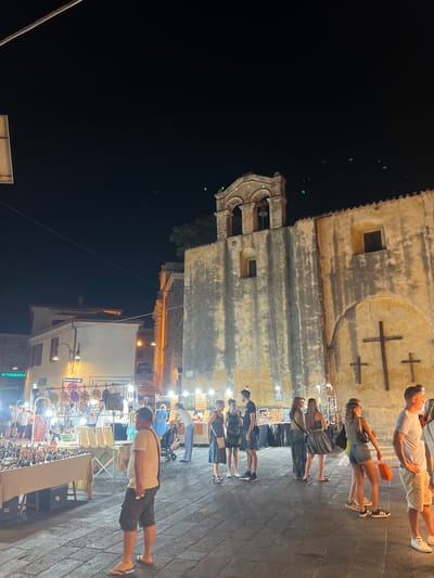 In the city of Orosei, Italy, at night, a group of people are gathered in a cobblestone plaza in front of an old stone building with a bell tower. The building has a weathered facade and a large arched entrance with three wooden crosses mounted on it. Several stalls are set up in the plaza, displaying various items for sale, including jewelry, bags, and trinkets. People are browsing the stalls and socializing. The scene is illuminated by artificial lights, creating a warm and inviting atmosphere. The sky is dark, indicating it is late at night.