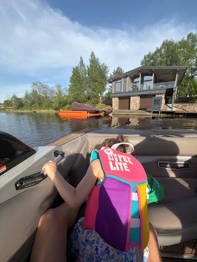 A girl in a colorful life vest is seated in a Centurion boat on a lake. The boat is near a lake house with a boat lift. The location may be Терехово, Russia.