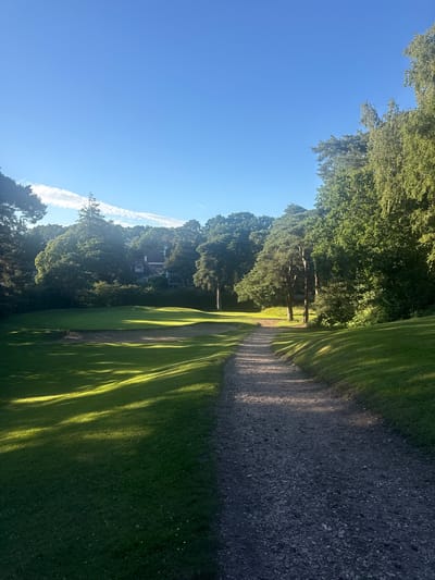 An upward-angle, vertical shot captures a sunlit golf course in Bournemouth, United Kingdom. The foreground features a gravel path curving to the right, bordered by lush green grass with distinct shadows cast by unseen trees. Beyond the path, the golf course unfolds with manicured fairways and sand traps. In the mid-ground, a cluster of mature trees partially obscures a red-roofed building. The background is dominated by a clear blue sky with a few wispy white clouds. The lighting suggests it is late afternoon, with the sun casting long shadows across the landscape. No people are visible in the scene.