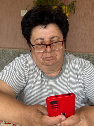 An elderly woman is holding a red smartphone in her right hand. She is wearing glasses and a striped shirt. She appears to be indoors, sitting on what looks like a granite countertop. In the background is a wall. The location is unknown.