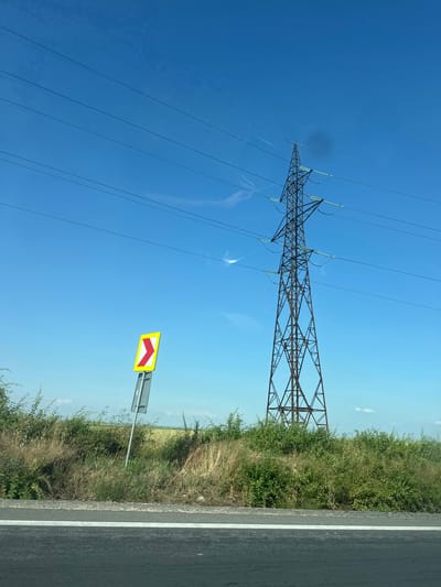 The image captures a rural roadside scene under a clear blue sky, likely taken from a vehicle in Moreni, Romania. Dominating the left side of the frame is a large, tall electricity pylon constructed from a metal lattice-work, supporting multiple power lines with visible insulators. These power lines extend across the bright sky, which is a deep, cloudless blue. A dark circular smudge or lens flare is noticeable in the upper-left sky area, along with some fainter reflections.
On the right side of the image, a section of an asphalt road is visible, marked with a clear white solid line along its edge. Bordering the road is a strip of dense vegetation, comprising both lush green bushes and drier grasses, suggesting an unkempt roadside area or natural growth. Amidst this foliage, a yellow rectangular road sign is mounted on a grey pole. The sign features two red chevron symbols pointing diagonally downwards, indicating a sharp bend or hazard.
The scene is brightly lit, characteristic of a clear day with sunny weather. No people or specific activities are depicted, but the perspective suggests the photo was taken while traveling. There is no visible text on the sign or elsewhere in the image. The general infrastructure, including the pylon and road sign design, is consistent with a European setting.