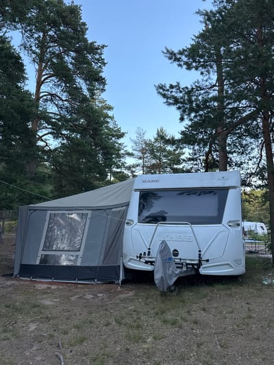 In Shepelevo, Russia, a white caravan with a dark grey annex is parked on a grassy area with pine trees in the background. The caravan has a large tinted rear window and a bike rack with a grey cover attached. The annex is connected to the caravan and has a plastic window. The sky is clear and blue, indicating it is daytime. The ground is covered with grass and pine needles. There are no people visible in the image.