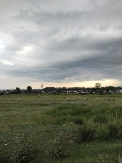 This is a landscape photograph taken in Московское, Russia, likely during the late afternoon or early evening. The sky is dramatically overcast with thick, dark grey clouds, suggesting the possibility of rain or an approaching storm.
The scene depicts a rural village nestled against a backdrop of trees. In the foreground, there is a field of tall green grass, with some sparse bushes and rocks scattered throughout. A dirt path or track runs horizontally through the middle ground of the field.
Dominating the mid-ground are utility poles with wires, indicating the presence of electricity or communication infrastructure in the village. The village itself consists of scattered houses with visible roofs, some appearing to be made of metal or tile. Wooden fences surround some of the properties.
In the distance, the trees form a dense line, and beyond them, the landscape appears to open up, possibly towards a horizon or a more distant plain. There are no people visible in the frame, and the overall mood is somewhat somber due to the heavy cloud cover. The lighting is subdued, contributing to the atmospheric, potentially moody feel of the scene. The time of day is suggested by the quality of light filtering through the clouds, indicating it's not midday. There is no visible text in the image.