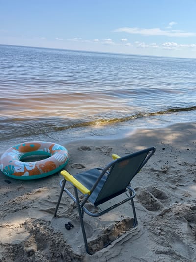 A beach chair and a colorful inner tube sit on a sandy shore beside calm, rippling water. The scene is likely a daytime beach setting, as suggested by the clear sky and bright light casting shadows. The water appears to be a lake or river, with gentle waves lapping at the sand. The sand is textured with footprints, indicating previous activity on the beach. A pair of small, dark flip-flops are visible near the chair's legs. The inner tube has a tropical pattern with orange and teal accents, featuring floral motifs. The beach chair is a portable, foldable type with a dark fabric seat and bright yellow armrests. The overall mood is relaxed and suggestive of a leisurely day by the water, perhaps for swimming or sunbathing. No people are visible in the frame. The sky is a pale blue with scattered white clouds. The sun is casting a warm glow on the water.