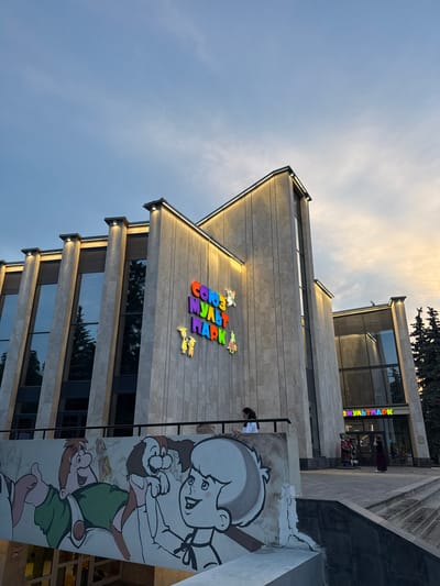 The image shows the facade of a building with a sign that reads "СОЮЗ МУЛЬТПАРК" in colorful, stylized letters. The building is modern, with large windows and a light-colored stone exterior. There are vertical lights illuminating the facade. In the foreground, there is a mural depicting cartoon characters. Two people are visible on the stairs leading to the entrance of the building, and one person is standing on a balcony to the left. Trees are visible to the right of the building. The sky above is a soft blue with some clouds, suggesting it might be dusk. The location is identified as Veshki, Russia.