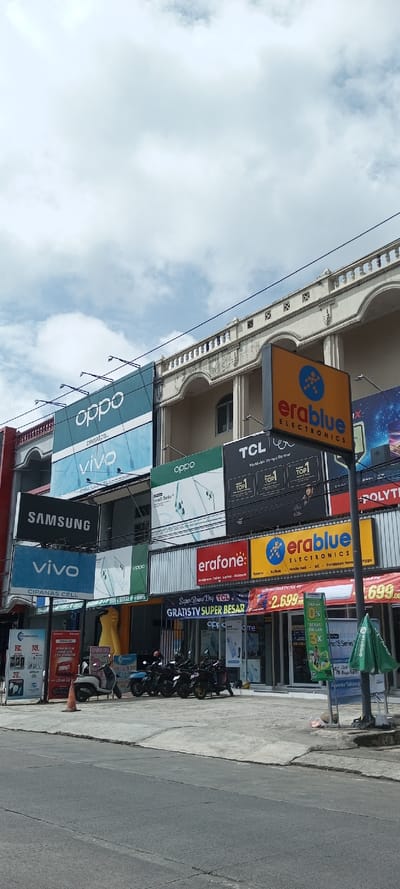 A multi-story building showcases a variety of electronics brands. Prominent signs for Oppo, Vivo, Samsung, TCL, and Polytron are visible. Several storefronts, including those for Erablue Electronics and Erafone, indicate retail activity. Motorcycles are parked in front of the stores. Signage advertises promotions like free televisions and special offers. The sky is partly cloudy, and a street runs in front of the building.