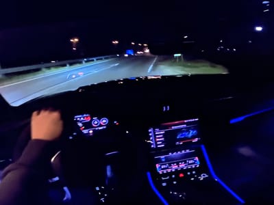 The image captures a first-person perspective from inside a modern car at night, likely on a highway. The dashboard displays are illuminated with blue and red lighting, indicating the car is operational. A hand is visible on the steering wheel, suggesting the driver is actively controlling the vehicle. The road ahead is dimly lit by headlights and streetlights, with the typical signs and guardrails of a highway visible. The car's interior ambient lighting is a prominent feature, casting a blue glow. The scene does not provide specific visual cues to confirm the location as Bezden, Bulgaria, but it represents a common scenario of nighttime driving.