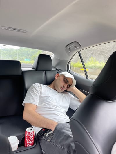 A man is asleep in the back seat of a car. He is lying down with his head near the rear passenger-side window, wearing a white t-shirt and a white baseball cap. A partially visible Coca-Cola can is in the armrest's cupholder next to him. The car appears to be in motion, and the view out the window shows a street sign that appears to read "Achara-".