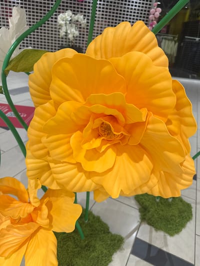 A large, vibrant orange flower, likely artificial, dominates the foreground of this image. Its petals are layered and textured, with a rich, saturated color. The flower appears to be part of a larger display, as green stems are visible, and other floral elements, including a white flower and another smaller orange flower, are partially in view. The background shows a checkered white wall, and behind it, out-of-focus elements suggest an indoor setting, possibly a retail or public space, with displays of clothing and possibly balloons visible. The floor is tiled with a pattern of light and dark grey squares. Two small patches of green, moss-like material are positioned at the base of the flowers, adding to the decorative arrangement. The image is captured from a close-up perspective, emphasizing the intricate details of the large orange flower. The location in Slitu, Norway is not indicated by any visible elements within the photograph.