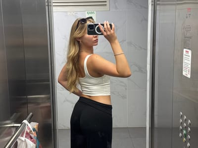 A woman with blonde hair, wearing a white crop top and black pants, takes a mirror selfie in an elevator. She is holding a smartphone in her right hand, with a magnetic phone ring attached. She has sunglasses perched on top of her head and wears a gold necklace and a silver bracelet. The elevator has a stainless steel finish and marbled tile walls. A shopping bag hangs from a handrail inside the elevator. The scene appears to be in a residential building in Ariel, Palestinian Territories.