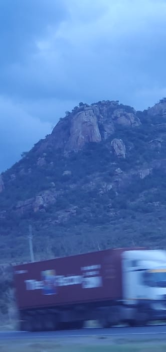 The image captures a vast, rocky mountain landscape in Kinanie ward, Kenya, under an overcast, blue-grey sky. The mountain, dominant in the background, is characterized by large, exposed granite formations interspersed with patches of dark green scrub and sparse vegetation. In the lower foreground, a white semi-trailer truck with a maroon-colored cargo container is traveling from left to right on a paved road. The truck and its container display significant motion blur, indicating rapid movement. Partial branding on the container includes the text "The Falcon" and "OM." No individuals are distinctly visible, though the truck's operation implies a driver. A utility pole is faintly visible on the left side, paralleling the road.