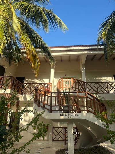An exterior view captures the "AILA HOUSE 09" guesthouse in Himmafushi, Maldives, under clear blue skies with strong sunlight. The two-story building features light-colored, possibly beige or off-white, walls and a flat or low-pitched roof with dark brown trim. Dominating the foreground is a light-colored, concrete spiral staircase with dark brown wooden railings, some exhibiting a diagonal lattice pattern. This staircase ascends to an upper-level balcony or walkway, which also has identical wooden railings.
On the first-floor landing of the spiral staircase, a large dark grey pot contains a green leafy plant. Another section of stairs with similar railings is visible to the left. A red fire extinguisher is mounted on the wall to the left of the spiral staircase. Entrances to rooms, some with glass panels and wooden frames, are visible on both the ground and upper floors. The ground area consists of light-colored paving stones or gravel with scattered green foliage and darker ground cover plants. Overhead, and partially obscuring the top of the building, are vibrant green palm fronds, casting distinct shadows on the building's facade. No individuals or active interactions are present in the scene.