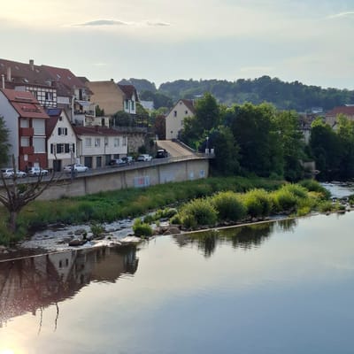 This media file depicts a scene in Gernsbach, Germany, likely during the late afternoon or early evening, given the soft, warm sunlight reflecting on the water and illuminating the sky with a pale haze. The image is oriented vertically, with the river and its reflection dominating the right side of the frame.
The foreground and middle ground feature a built-up area along the riverbank. Buildings with red-tiled roofs and white or light-colored facades line a street. Some buildings display traditional half-timbered architectural elements. Several cars are parked along the street, indicating an inhabited area. A concrete embankment with grassy vegetation separates the street from the river. The river itself is calm, with gentle ripples creating a mirror-like reflection of the buildings, trees, and sky.
Beyond the buildings, a densely wooded hill rises, covered in lush green trees. The trees are vibrant, suggesting a summer or late spring setting. The sky is a pale blue with some wispy clouds, and the sun is a bright, hazy orb low on the horizon, further suggesting the time of day.
The overall mood is tranquil and picturesque. The reflection on the water adds a layer of depth and beauty to the scene. There are no people visible in the image. Some graffiti is present on the concrete embankment. The specific location cues, such as the architectural style of the buildings and the presence of the river, are consistent with a European town.