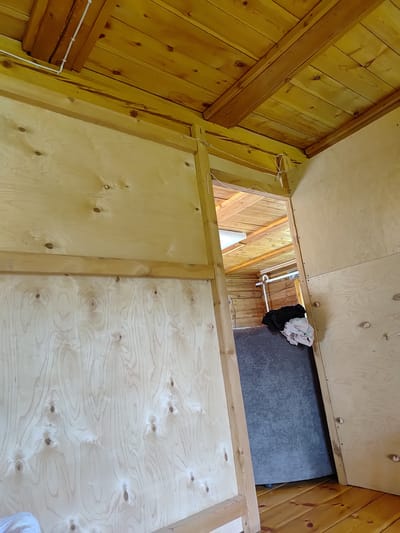 The image shows the interior of a rustic wooden structure, possibly a shed or cabin, with unfinished plywood walls and a wooden plank ceiling supported by beams. A doorway in the center leads to another section of the structure, revealing more wooden walls and ceiling. On the floor of this second area, a large, dark, cylindrical object covered with fabric is visible, along with a white pipe attached to a wall. There are no people or animals present in the image. The scene appears to be located in a rural setting, consistent with a location in the village of Baryntsy, Russia. The lighting suggests it is daytime, with natural light entering from an unseen source.