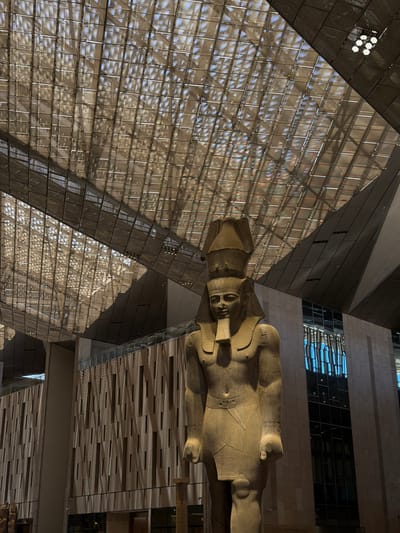 The image shows a large ancient Egyptian statue of a pharaoh, likely Ramses II, positioned in the grand atrium of the Grand Egyptian Museum in Kafrat Nassar, Egypt. The statue, made of granite, stands prominently in the foreground, with its intricate details and regal headdress clearly visible. It is illuminated by natural light filtering through the museum's impressive, geometrically patterned glass roof, which casts dappled shadows onto the surrounding architecture.

The museum's interior features modern architectural elements, including a facade of stacked rectangular panels and large glass windows that reflect the sky. In the background, a contemporary building rises, contrasting with the ancient statue. The lighting is a mix of natural sunlight and strategically placed spotlights on the ceiling. There are no people visible in the immediate vicinity of the statue, emphasizing its solitary grandeur within the vast museum space. The scene conveys a sense of awe and historical significance, blending ancient heritage with modern design.