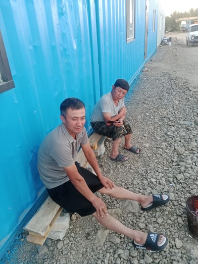 Two adult males are seated on a wooden plank outside a blue prefabricated structure. The man in the foreground is wearing a grey polo shirt and black shorts, and has his legs extended. He is looking towards the viewer with his arms resting on his knees. The second man, seated to his right, is wearing a grey t-shirt and camouflage shorts, and is holding a mobile phone. He is looking away from the viewer. The ground is covered in gravel. In the background, there are several vehicles, including a white SUV and a truck. The scene appears to be in an outdoor industrial or construction area.