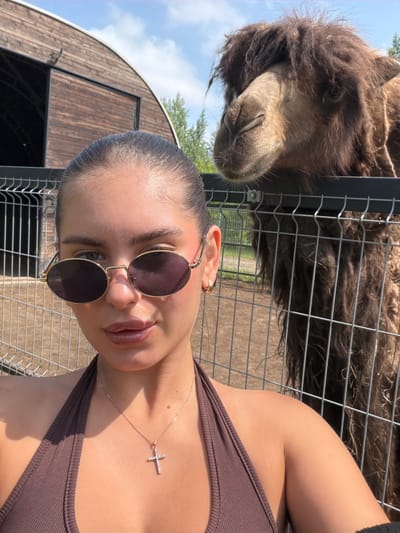 A young woman with dark hair pulled back is taking a selfie. She is wearing sunglasses and a brown halter top with a cross necklace. Behind her, a camel is visible, with its head over a fence. The setting appears to be an outdoor enclosure, possibly a zoo or sanctuary, with a wooden building and trees in the background. The lighting suggests it is daytime, with the sun casting shadows.
