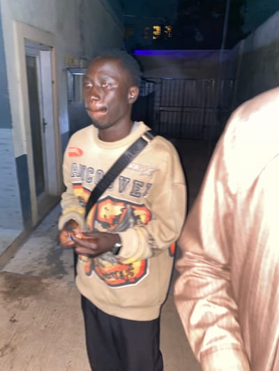 A young Black male stands outdoors at night in what appears to be an alleyway in Jos, Nigeria. He is wearing a light brown sweatshirt with a graphic design featuring text and a fiery image. A black strap from a bag crosses his chest. His hands are clasped in front of him, and he appears to be holding something small. His face is turned towards the camera, and he has a neutral expression. To his right, a portion of another person's shoulder and arm is visible, wearing a shiny, light-colored garment. The background shows a textured wall on the left and a dark fence with vertical slats on the right. Above the fence, some faint blue light is visible. The ground is paved with concrete. The overall lighting suggests it is nighttime.