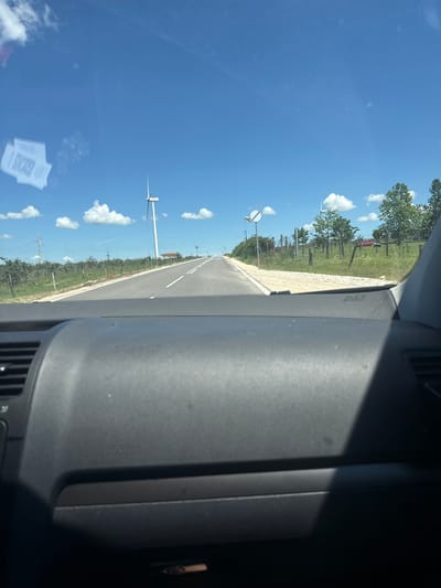 The image shows a view from inside a car traveling on a paved road. The road is lined with sparse vegetation and some trees on either side. In the distance, a large wind turbine is visible against a bright blue sky with scattered white clouds. There are also some power lines and a small red structure with a roof visible on the right side of the road. The dashboard of the car is in the foreground, with a visible "AIRBAG" label on it. On the left side of the windshield, several QR codes are affixed. The scene appears to be a rural or semi-rural landscape.
