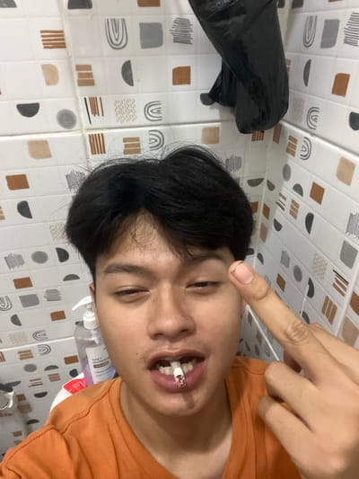 A young man is taking a selfie in a bathroom with patterned tiles. He has dark hair and is wearing an orange t-shirt. He is holding a lit cigarette in his mouth and is gesturing with his right middle finger. A black trash bag is hanging on the wall behind him, and a bottle of hand soap is visible on a shelf. The man appears to be in a bathroom in Serdang Kulon, Indonesia.