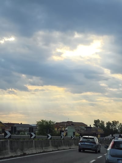 The image shows a view from a vehicle traveling on a highway in Casaluce, Italy. The sky is filled with dramatic clouds, with patches of sunlight breaking through, creating a dramatic lighting effect. The highway has a concrete barrier with black and white chevron directional signs. Beyond the barrier, there are residential buildings, some with visible solar panels, and trees. Two cars are visible on the highway; the one in the foreground is a dark grey station wagon with the license plate CZ 325MX, and another car is partially visible to its right. The overall scene depicts a typical highway landscape with a backdrop of Italian architecture and a dynamic sky.