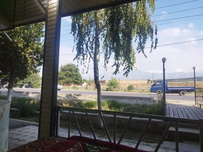 This image depicts an outdoor scene, possibly a covered patio or veranda, with a view of a street and distant mountains. In the foreground, a railing made of metal bars runs horizontally, supported by diagonal struts. The floor beneath the railing appears to be paved with patterned tiles. A patterned blanket or rug is visible on the ground in the lower left corner.
Beyond the railing, a tree with long, drooping branches covered in green leaves dominates the middle ground, casting shadows. To the left of this tree, another tree with a white-barked trunk is visible. A paved road runs horizontally across the scene, with a light-colored sedan parked on the side and a blue pickup truck driving away from the viewer. Green grass and vegetation line the roadside.
In the background, a brick wall runs along the edge of the road, and behind it are more trees and some buildings. Distant mountains form the horizon under a bright, cloudy sky with visible power lines stretching across it. Two streetlights with spherical tops are also present in the mid-ground, to the right of the blue truck. The overall impression is a daytime scene in a semi-urban or suburban setting, characterized by natural elements like trees and mountains, juxtaposed with man-made structures and vehicles. The location is identified as Kyzyl-Kyya, Kyrgyzstan.