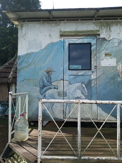 An old, weathered building features a mural of a cowboy riding a horse against a mountain backdrop. A door with a small window is painted to blend with the artwork. A white, rusty metal railing surrounds a wooden deck in front of the building. A large, clear water bottle with a red cap sits on the deck next to the railing. The building's exterior shows signs of age and wear.
