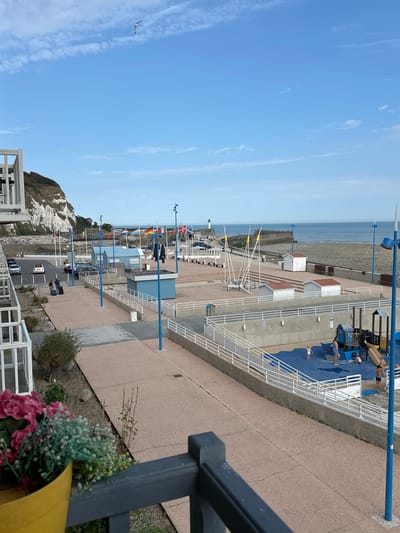 The image shows a view of the promenade and beach area in Saint-Valery-en-Caux, France, on a clear day. In the foreground, a yellow pot with pink and white flowers sits on a dark railing. The promenade, paved with reddish-brown aggregate, stretches towards the background. To the left, a steep, white-cliffed hill rises above a parking lot with several cars. A few people are walking with luggage on the promenade.
Further back, a playground with blue surfacing and various play structures, including slides and swings, is visible. Several small, white beach huts with red roofs are lined up along the promenade. Beyond the huts, a sandy beach meets the blue ocean, with a harbor wall and a lighthouse in the distance. Various national flags are displayed on flagpoles along the promenade. The sky is blue with scattered white clouds, and a seagull is flying high above.