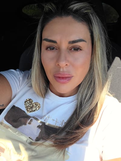 The image is a close-up, frontal, selfie-style shot of a woman sitting in what appears to be a car. The woman has blonde and brown highlighted hair, styled long and parted to her left. She is wearing a white t-shirt with a large, shimmering gold heart graphic and a brown, abstract print. Sunlight streams in from the right side of the frame, casting shadows on her face and the car's interior, which is partially visible as textured gray fabric on the right. Her makeup is visible, with emphasis on her eyes and lips. The background is dark, suggesting the interior of a vehicle, with a hint of green foliage visible through a window on the upper left. The image does not provide any specific details that would place it in the city of Ravnogor, Bulgaria.