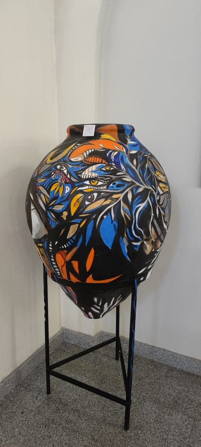 An ornate, black ceramic vase, richly decorated with abstract patterns in orange, blue, yellow, and white, is displayed on a black metal stand. The vase has a globular shape with a narrow neck. The intricate artwork features eye-like motifs and swirling organic forms. A small QR code sticker is attached to the rim of the vase. The stand is a simple, industrial design with four legs forming an X-shape at the base. The vase and stand are positioned in the corner of a room with light-colored walls and a speckled grey tiled floor. The scene is lit with neutral lighting, casting soft shadows. The overall impression is of an art display. Based on available metadata, this image was taken in Voskevaz, Armenia.