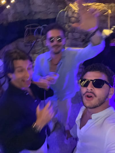 Three adult males are captured in a dimly lit, close-up photograph taken in Poltu Quatu, Italy. The environment features textured, rock-like walls illuminated predominantly by purple and blue light, with accents of warm yellow overhead. Motion blur is evident across the image, particularly on the subjects' limbs and faces, indicating movement during exposure.

In the foreground, on the right, a man with dark, curly hair, a mustache, and a short beard is wearing black sunglasses that reflect distorted light patterns. He wears a white, open-collared shirt and has his mouth open in an "O" shape, looking directly towards the camera.

Positioned behind and slightly to the left of the foreground subject is a man wearing a light blue collared shirt and light-colored pants. He has a mustache and short beard, and his eyes are obscured by dark sunglasses. He holds a clear glass or plastic cup containing a light-colored beverage and ice in his left hand. His right arm is raised high and heavily blurred, suggesting an active waving or gesturing motion.

On the far left, partially visible, is a third man with dark hair, wearing a dark polo shirt. His face is motion-blurred, but an open-mouthed expression is discernible. A watch or bracelet is visible on his left wrist, which is also blurred. The men appear to be in close proximity, suggesting a social gathering.