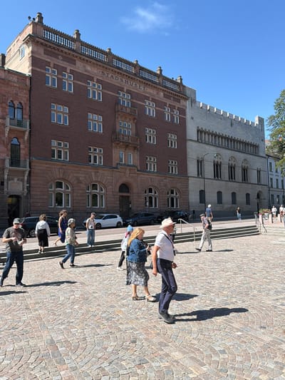 The image depicts a sunny day in Lund, Sweden, with a clear blue sky overhead. In the background, the prominent Stadshuset (City Hall) building dominates the scene. The building features a reddish-brown brick facade with intricate architectural details, including arched windows and balconies. To the right of the Stadshuset, a lighter colored building with a more modern, fortress-like appearance stands. In the foreground, a cobblestone plaza is visible, with several people walking across it. Some individuals are captured mid-stride, while others are engaged in conversation. Cars are parked along the street behind the plaza, and some foliage can be seen on the right side of the image. The overall impression is of a public square in a European city, bustling with activity on a pleasant day.