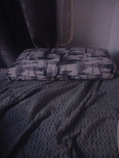 A pillow with a black and white abstract pattern rests on a bed covered with a dark blue textured blanket. The pillow is positioned towards the center of the bed, with the blanket showing rippling folds. In the background, dark curtains are visible, and a sliver of a light-colored wall can be seen. The lighting is dim, casting shadows and creating a subdued atmosphere. The image appears to be taken indoors, likely in a bedroom. The location in Sibircevo, Russia, is not discernible from the visual information present in the image.