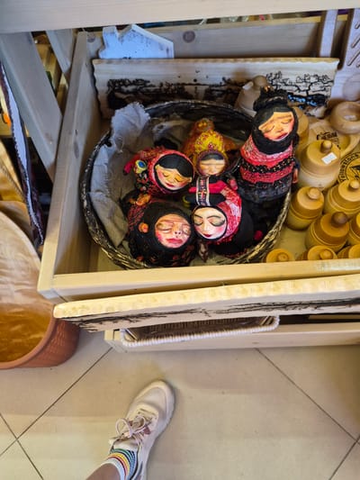 The image depicts a close-up view of a wooden shelf unit filled with various handcrafted items. In the upper section of the shelf, a woven basket contains several Matryoshka dolls. These dolls are adorned with traditional attire, featuring floral patterns and scarves. Some dolls have painted faces with closed eyes and rosy cheeks, while others are partially obscured. To the right of the basket, several wooden containers with lids are stacked, some with visible labels. Below the basket, a decorative wooden plaque with a carved silhouette of trees is visible. Further down, another shelf holds a woven basket. The overall environment appears to be a shop or market stall. In the foreground, the foot of a person wearing a white sneaker and striped socks is visible, suggesting the perspective of someone browsing the merchandise. The floor is tiled in a beige color with dark grout lines. The scene is lit with natural or soft artificial light, creating a warm and inviting atmosphere. This scene is likely from the city of Krasnyy Klyuch, Russia, given the presence of traditional Russian dolls.