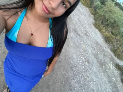 A young woman with dark hair and fair skin is standing outdoors, possibly on a dirt path or a dry, rocky incline. She is wearing a blue and turquoise halter-neck dress and a gold necklace with a rose pendant. Her gaze is directed towards the camera, and she has a slight smile. The background features greenery and a sparsely vegetated area. The lighting suggests it is daytime. There are no discernible landmarks within the image that would specifically place her in the city of Vera, Spain.