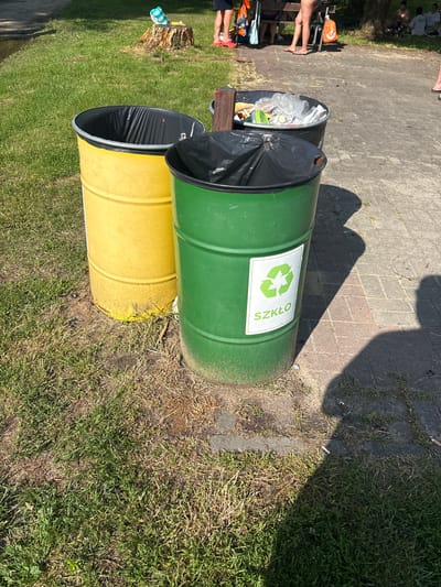 This is a medium-angle, portrait-oriented shot of two large, horizontal recycling bins placed on a grassy area next to a paved walkway. The bin on top is green and the one below it is yellow. Both bins have black liners visible inside them. The green bin has a white sticker with a green recycling symbol and the word "SZKŁO" which means "glass" in Polish. The scene appears to be outdoors on a sunny day, indicated by the shadows cast on the ground.
In the background, on the paved walkway, several people are sitting and standing. Some are wearing casual clothing. One person is partially visible at the top left of the image. The overall setting suggests a public outdoor space, possibly a park or recreational area. The grass is green and well-maintained, with some patches of dry grass or dirt. The paved area seems to be made of interlocking concrete pavers. The presence of the recycling bins and people suggests a gathering or a place where people are enjoying an outdoor activity. The lighting and shadows indicate it is likely midday.
