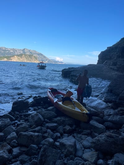 Two individuals are standing near two kayaks on a rocky shore, in what appears to be a cave or overhang. One person is wearing red swim trunks and is holding a phone. The other person is wearing blue shorts and has a blue backpack slung over their shoulder. A yellow kayak is positioned in front of them, and a red kayak is partially visible behind it. The kayaks are on the water, which is choppy, suggesting an active environment. In the background, mountains rise dramatically from the water, with some vegetation on their slopes. A boat with several people on board is also visible on the water, further in the distance. The sky is a clear, bright blue, indicating a sunny day. The time of day appears to be midday, given the bright sunlight and clear sky. The overall mood seems to be relaxed and adventurous, as people prepare for or have just finished an activity on the water. There are no visible signs or text in the image. The scene is set by a rocky shore and a body of water, likely a river or a bay, with mountainous terrain surrounding it. The presence of kayaks and a boat suggests a recreational or touring activity.